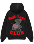 A black pullover hoodie shown from both the front and back. The front is plain, while the back features a graphic of a gray teddy bear holding a pink heart, surrounded by small rhinestone-like dots, with the words 'Self-Love CLUB' in red arched above and below.