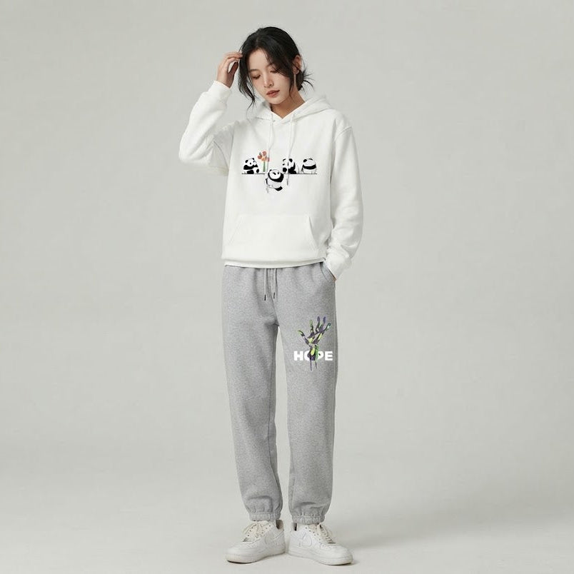 A model wearing a white hoodie with black Arabic calligraphy and colorful floral details, paired with light gray sweatpants featuring a small graphic and the word 'HOPE', on a light gray studio background.