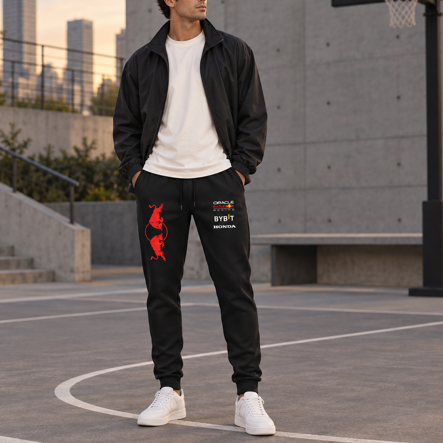 A man stands on an outdoor basketball court wearing a black zip-up jacket, white t-shirt, black sweatpants with red and white graphics, and white sneakers. The background shows a concrete wall, stairs, and a basketball hoop at sunset.