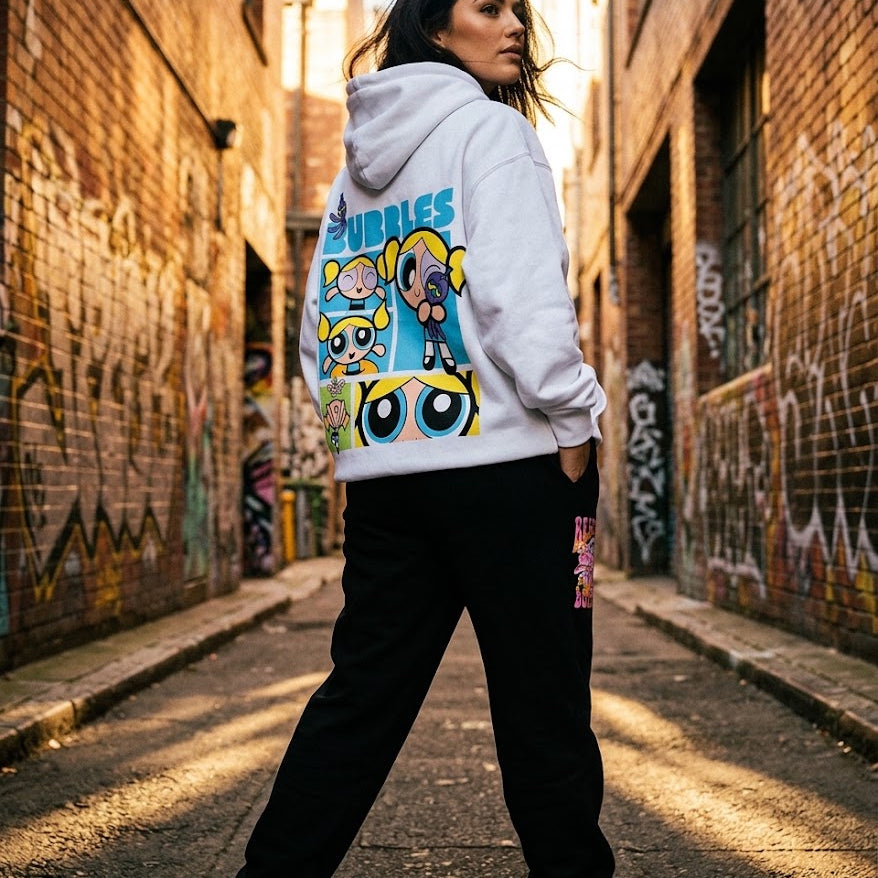 A woman stands in a sunlit urban alley wearing a white pullover hoodie with a large colorful Powerpuff Girls graphic on the back, paired with black sweatpants.