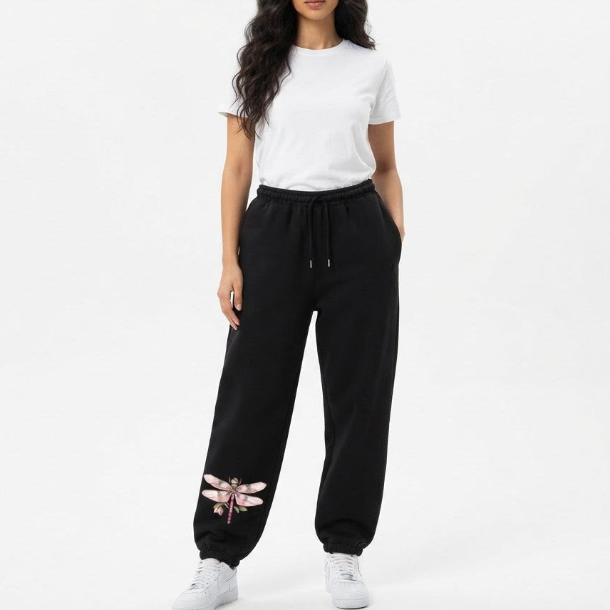 A woman modeling black sweatpants with a pink dragonfly graphic near the ankle, styled with a white t-shirt and white sneakers against a plain white studio background.